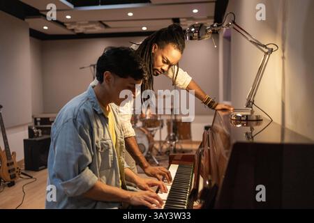 Two musicians work together at a piano, collaborating creatively in a modern recording studio. Instruments and equipment are in the background, highli Stock Photo
