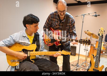In a recording studio, a guitar instructor guides a student, who is holding a guitar, through a session. Instruments and microphones are set up around Stock Photo