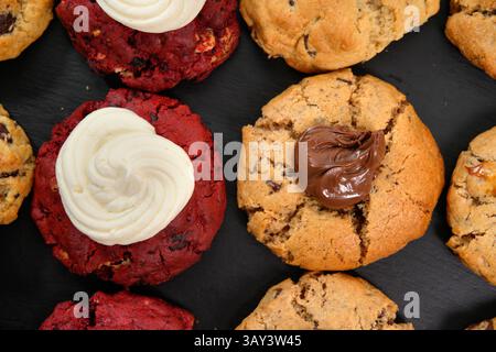 Variety of homemade cookies, including chocolate chip and red velvet topped with cream, displayed on a black surface. Stock Photo