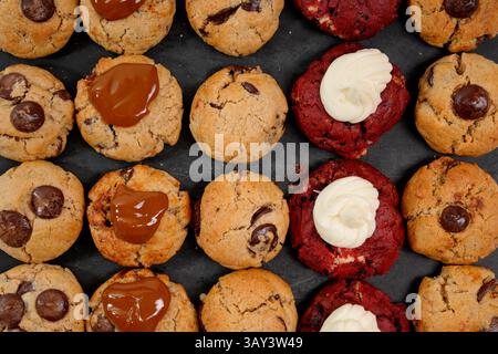 Top view of a variety of freshly baked cookies, including chocolate chip, red velvet, and caramel-topped, arranged neatly on a dark surface. Stock Photo