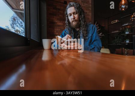 A bearded man with long hair sits at a wooden table near a window, writing in a notebook with a focused expression. A white mug adds warmth to the coz Stock Photo