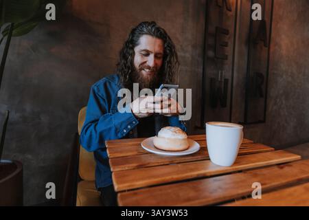 A man with long hair and a beard smiles while using his smartphone at a wooden table in a cozy cafe. A cup of coffee and a pastry are in front of him, Stock Photo