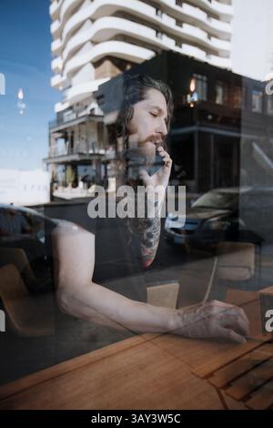 A reflective urban scene shows a man with tattoos, deep in thought, working on a laptop by the window of a modern building with unique architecture. T Stock Photo