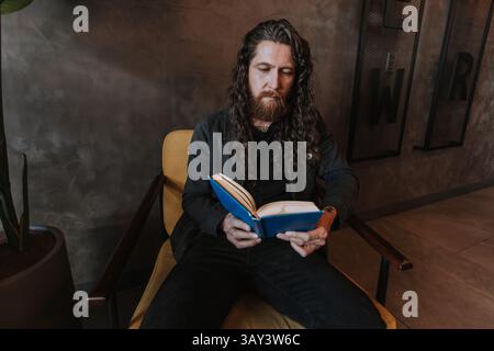 A man with long hair and a beard is sitting on a yellow chair reading a book with a blue cover, surrounded by modern decor, creating a relaxed and int Stock Photo