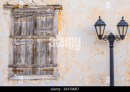 An old wooden window shutter and vintage street lamp on a rustic, weathered wall in Triovasalos, Milos Island, Greece, showcase the area's timeless ch Stock Photo