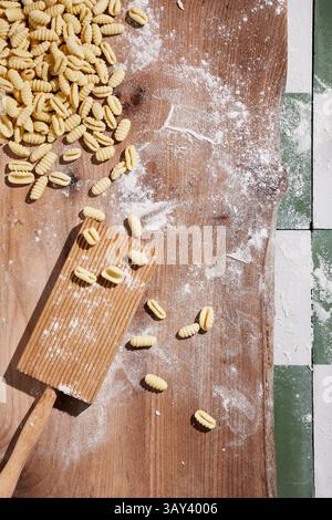 Top view of fresh homemade gnocchi scattered on a flour-dusted wooden surface beside a gnocchi board and rolling pin, highlighting the process of past Stock Photo