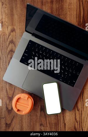 A top view of a laptop and a smartphone with a blank screen placed next to a thermos cup on a wooden surface, suggesting an organized remote workspace Stock Photo