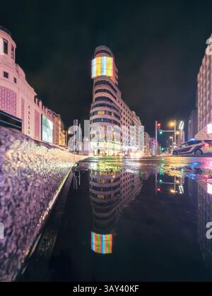 Vibrant nighttime cityscape of Madrid featuring a historic building with colorful lights, reflected in a puddle on Gran Via street. Urban ambiance is Stock Photo