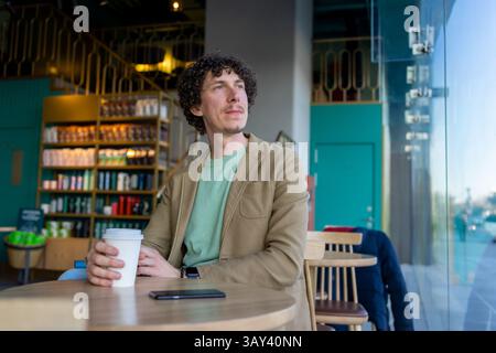 A thoughtful man sits in a modern cafe, holding a coffee cup and looking out the window. Bright interior, shelves full of products, and a relaxed atmo Stock Photo