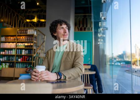 A businessman with curly hair sits at a cafe table with a coffee cup. He gazes outside through a large window, dressed casually in a green shirt and b Stock Photo