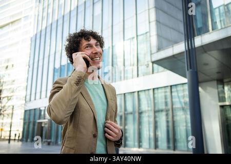 A cheerful man in a blazer is talking on his smartphone outside a sleek, glass paneled office building. The image captures urban business communicatio Stock Photo
