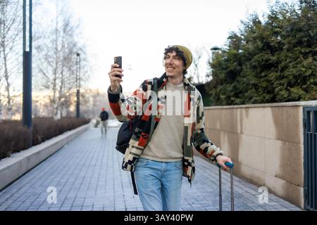 A cheerful man with curly hair and a colorful jacket smiles while taking a selfie on a bright city street. He holds a suitcase, capturing memories of Stock Photo