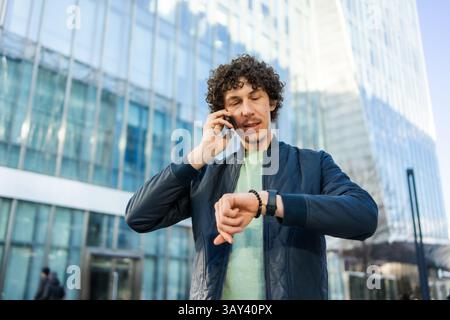 A businessman stands in front of a glass building, talking on his smartphone while checking the time on his smartwatch, symbolizing productivity and m Stock Photo