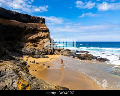 From above a lone woman walks on the sandy shore of Tindaya Beach in Fuerteventura, amidst rugged cliffs and foamy sea waves. Stock Photo