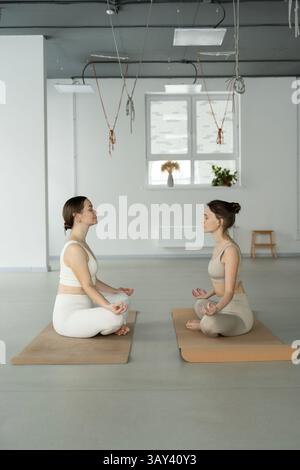 Two women sit cross legged on yoga mats, practicing mindfulness meditation in a serene studio setting. Natural light streams through the window, enhan Stock Photo