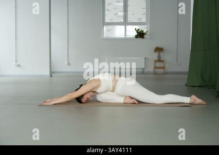 A woman practicing a deep stretch in a peaceful, minimalist studio. The quiet environment and natural light enhance the calming atmosphere of the yoga Stock Photo