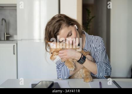 A woman embraces a ginger cat while sitting at a table in a cozy kitchen. She wears striped clothing and earbuds, exuding warmth and contentment in a Stock Photo