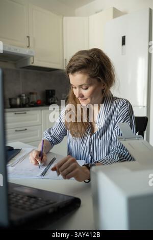 A woman in a striped shirt is focused on writing in a notebook while working from home in her kitchen. A laptop and other office items surround her on Stock Photo