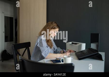 A woman in a striped shirt works at home, focusing on writing in a notebook beside a laptop. A cup of coffee and a printer are visible on the desk. Th Stock Photo