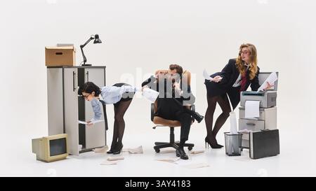 Two women bent over in workplace, handing papers to seated male coworker multitasking on phone near messy printer station. Stock Photo