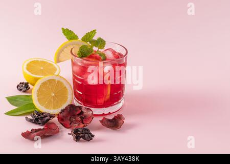 A vibrant red hibiscus drink with ice cubes, garnished with fresh lemon slices and mint leaves, surrounded by dried hibiscus petals on a soft pink bac Stock Photo