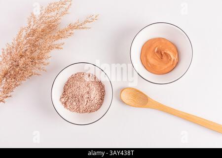 Top view of dry cosmetic clay powder and mixed clay facial mask in white bowls with wooden spoon and dried grass, isolated on a white surface. Stock Photo