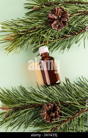 Top view of a brown glass bottle containing essential oil, situated among vibrant green pine needles and pine cones on a soft green background. Stock Photo