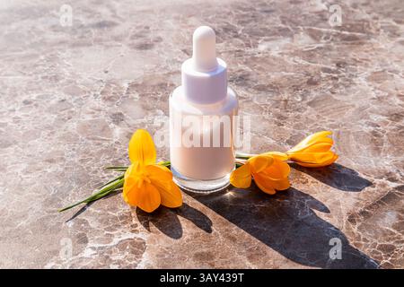 A white dropper bottle on marble, paired with bright yellow flowers. The sunlight enhances the contrast, creating a serene and elegant aesthetic suita Stock Photo