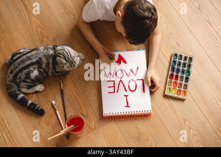 A boy lies on a wooden floor, painting a heartfelt Mother's Day card with I Love Mom. Nearby, a curious cat watches, surrounded by art supplies and br Stock Photo