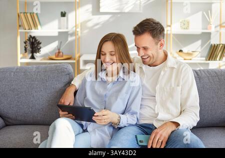 Excited cheerful young happy family couple using tablet computer, getting news sitting, chatting  Stock Photo