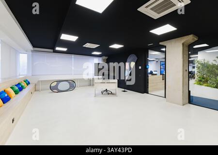 Modern and spacious training room featuring a long bench with colorful cushions, a large TV screen, blackboard, and bright white flooring. Stock Photo