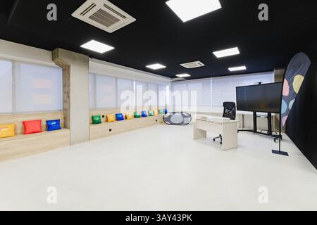 Modern and spacious training room featuring a long bench with colorful cushions, a large TV screen, blackboard, and bright white flooring. Stock Photo