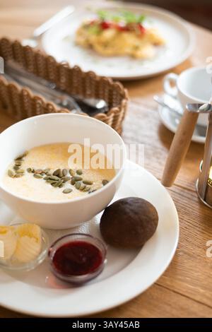 A cozy breakfast setting in a cafe featuring a bowl of creamy corn porridge garnished with seeds, accompanied by scrambled eggs, butter, jam, and a ru Stock Photo