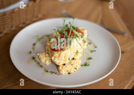Scrumptious breakfast of creamy scrambled eggs garnished with cherry tomatoes and greens, elegantly served on a white plate at a cozy cafe, perfect fo Stock Photo