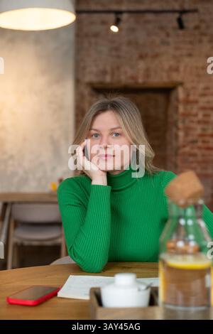 A woman in a green sweater sits at a cafe table, surrounded by a warm, cozy ambiance. With her hand resting on her face, she appears relaxed and thoug Stock Photo