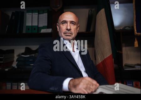 A confident businessman in a suit sits at a desk in an office setting. Shelves with binders and a flag in the background create a professional atmosph Stock Photo