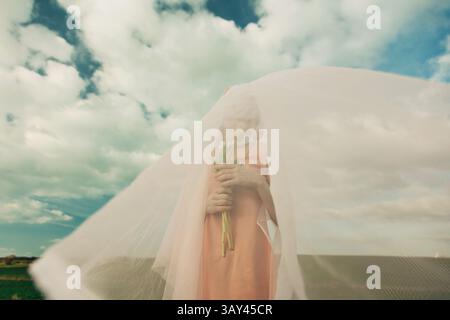 A woman stands under a translucent white veil, holding tulips. Her face is obscured, creating an ethereal and mysterious atmosphere against a backdrop Stock Photo