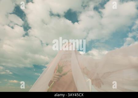 A woman draped in sheer fabric stands under a vibrant sky, creating an ethereal, dreamlike scene. The soft clouds provide a serene, mystical backdrop. Stock Photo