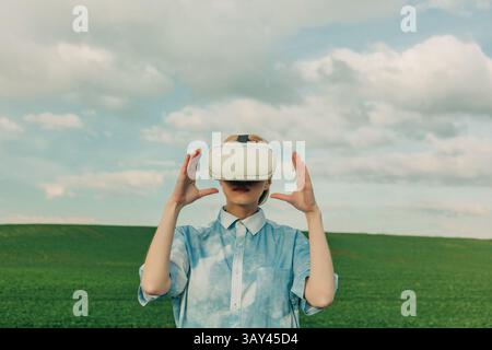 A woman enjoys virtual reality in a scenic outdoor setting, wearing a VR headset against a backdrop of green fields and a blue sky, blending technolog Stock Photo