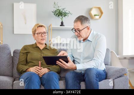 Senior couple sitting on sofa in living room at home and using tablet together browsing apps Stock Photo