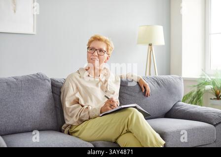 Blond woman sitting on gray sofa with notebook and pen, smiling and making notes in bright room. Stock Photo