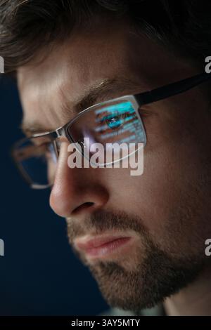 Close-up a focused data scientist wearing glasses, with lines of computer code reflecting on his lenses. Software engineer analysing data late in a dark room.  Stock Photo