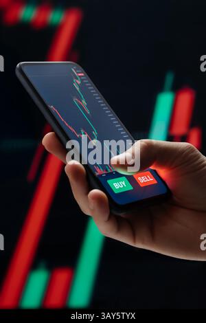 Hand holding smartphone with trading application on screen against background with crypto graphs. Close up of trader hand buying cryptocurrency online via stock market app on his mobile phone Stock Photo