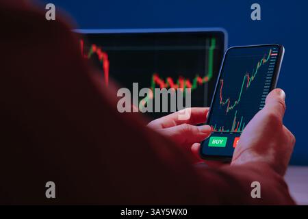 Trader hands holding smartphone, using trading app for analyzing financial, stock market and cryptocurrency data, with red and green candlestick charts on screen. Stock Photo