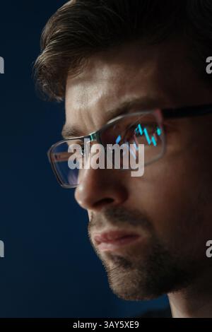 Close up, focused crypto trader wearing glasses closely analyzes cryptocurrency or stock market data with candlestick charts and financial graphs reflecting in his lenses.  Stock Photo