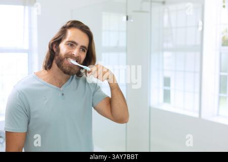 Young man brushing teeth in modern bathroom, with toothbrush and mirror, copy space Stock Photo
