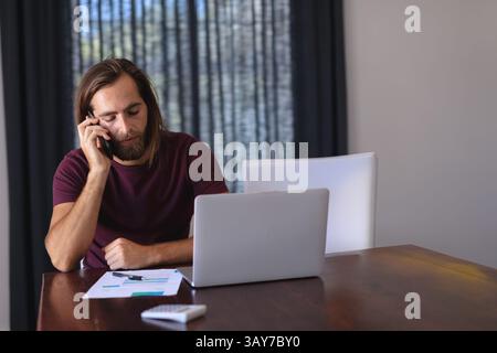 Young adult male talking on smartphone at home office table, with laptop, charts and calculator Stock Photo