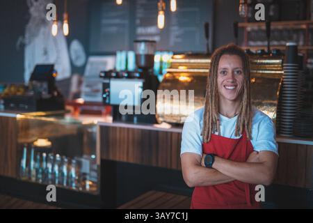 Young adult male barista standing smiling behind counter in cafe with steaming espresso machine Stock Photo