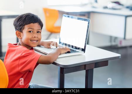 Boy typing on laptop at desk smiling at camera in classroom, with orange chairs, copy space Stock Photo