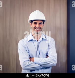 Man standing in site office wearing white hard hat and light blue shirt, smiling confidently Stock Photo